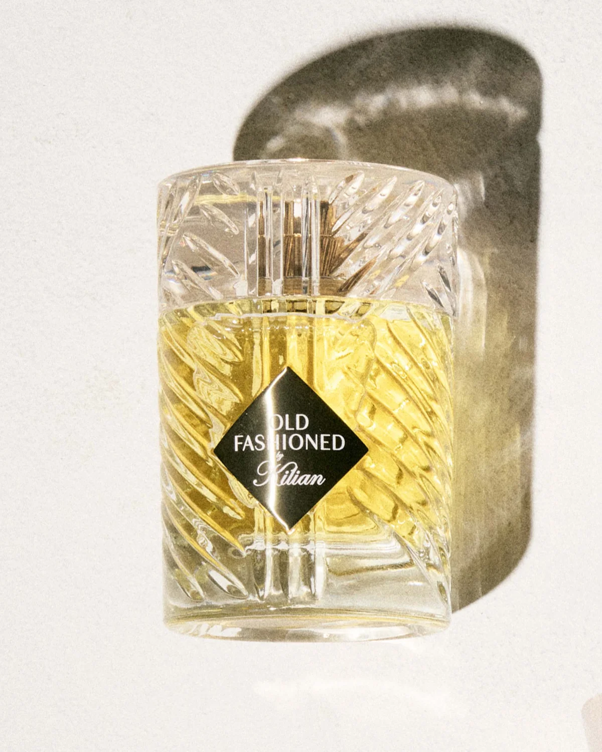 A clear perfume bottle labeled "Old Fashioned" by Kilian, with a shadow on a light textured surface. A clear perfume bottle labeled "Old Fashioned" by Kilian, with a shadow on a light textured surface.