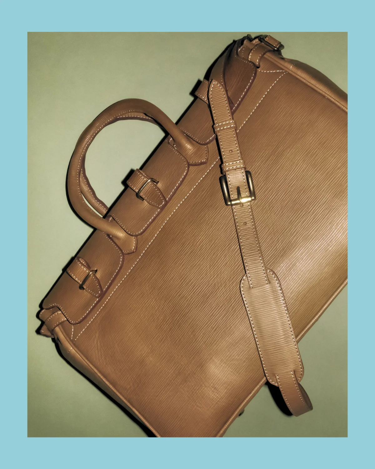 A sleek brown leather overnight bag with a shoulder strap against a light blue background.