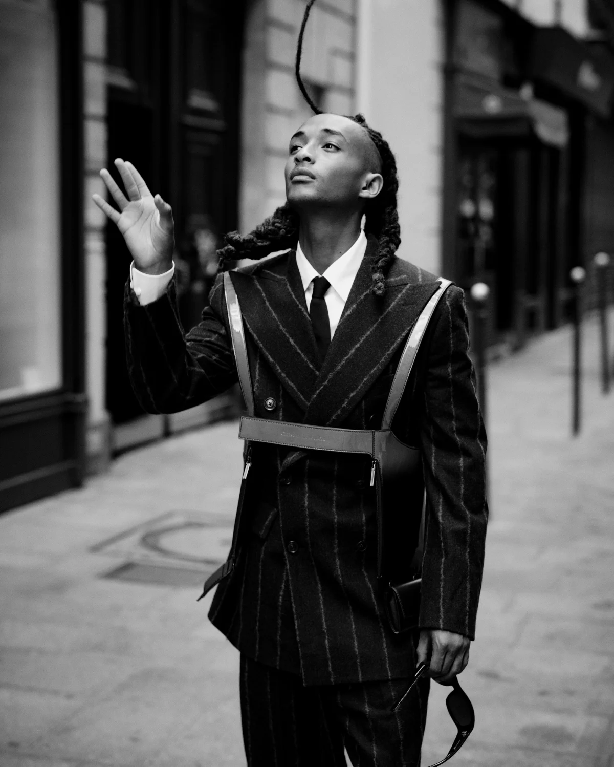 A person with braided hair, wearing a pinstripe suit, stands in a city street, gesturing with one hand.