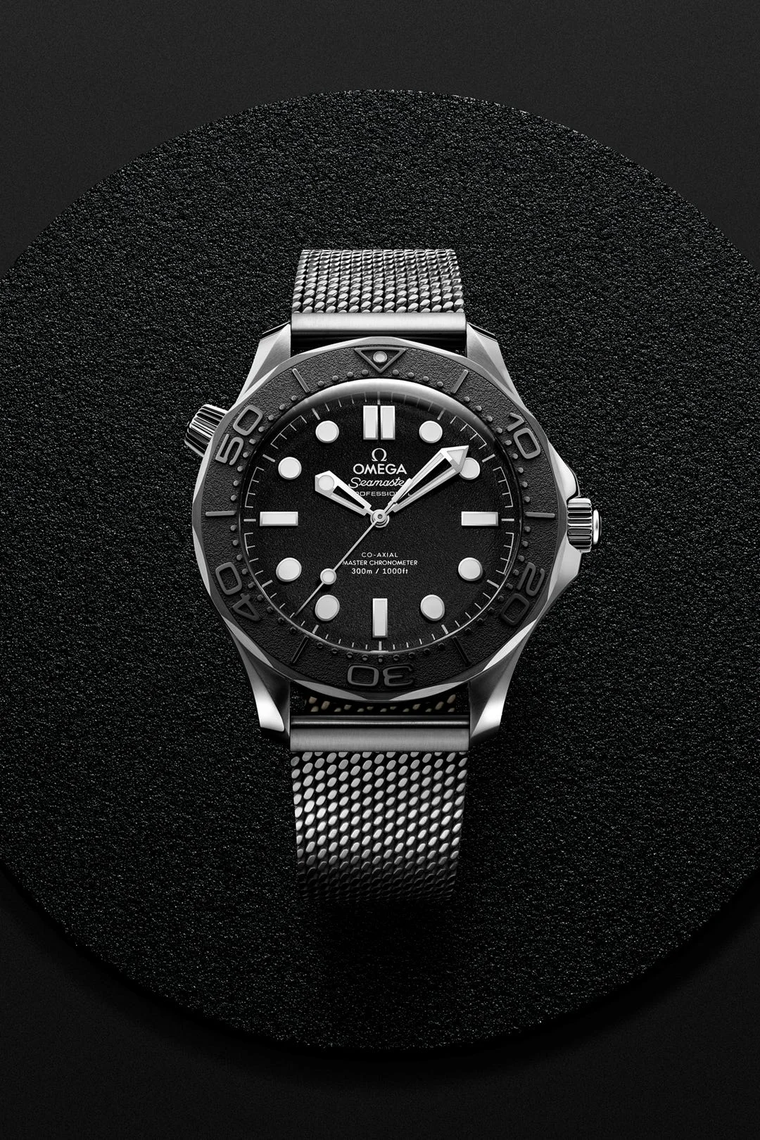 Black Omega Seamaster watch with silver mesh strap displayed on a textured round surface.
