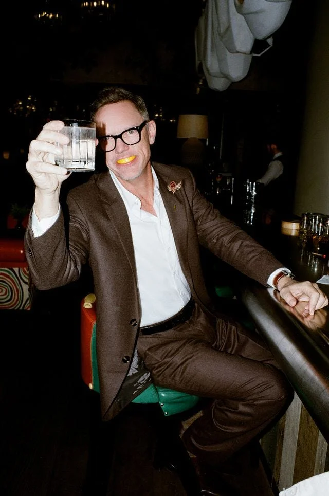 Person in a brown suit holding a glass and smiling, with a yellow object in their mouth, at a bar setting. Person in a brown suit holding a glass and smiling, with a yellow object in their mouth, at a bar setting.