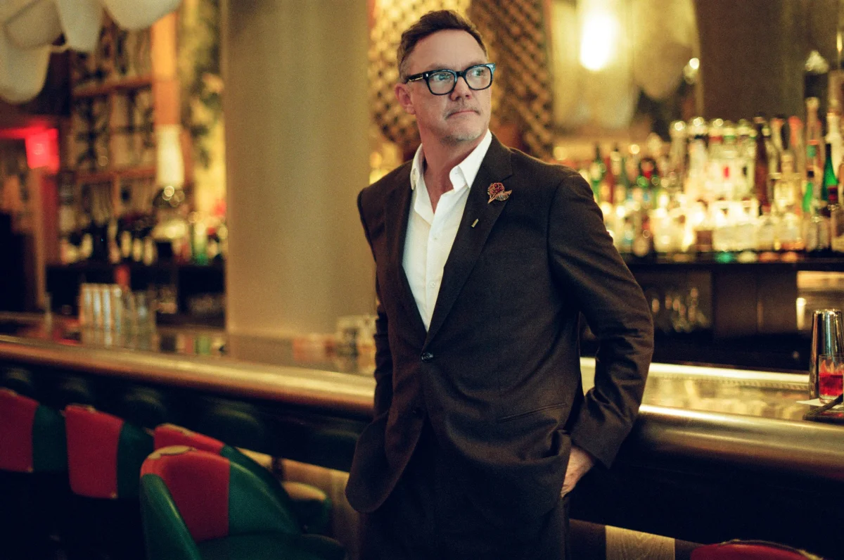 A man in a brown suit and glasses stands confidently at a bar, with colorful seating and a vibrant backdrop. A man in a brown suit and glasses stands confidently at a bar, with colorful seating and a vibrant backdrop.