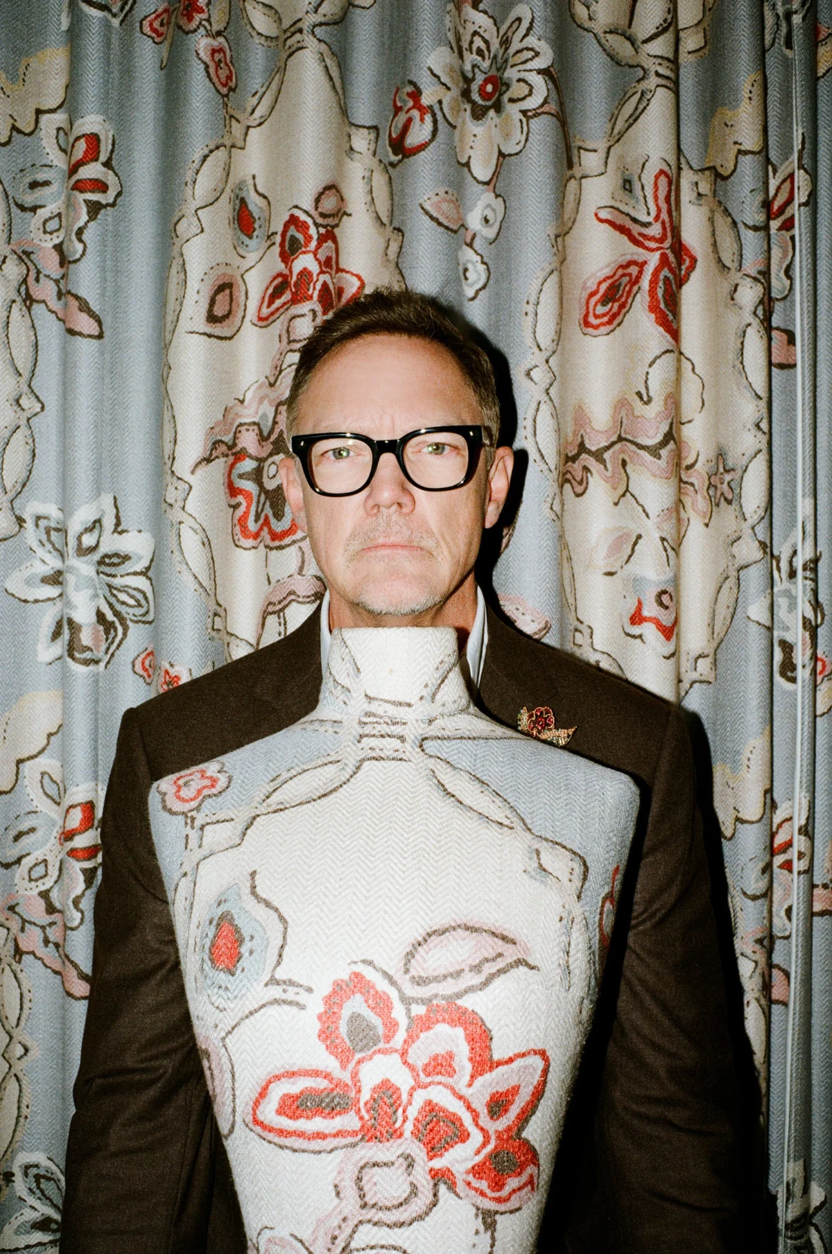 A person wearing glasses stands in front of a floral curtain, holding a decorative mannequin torso adorned with patterns. A person wearing glasses stands in front of a floral curtain, holding a decorative mannequin torso adorned with patterns.
